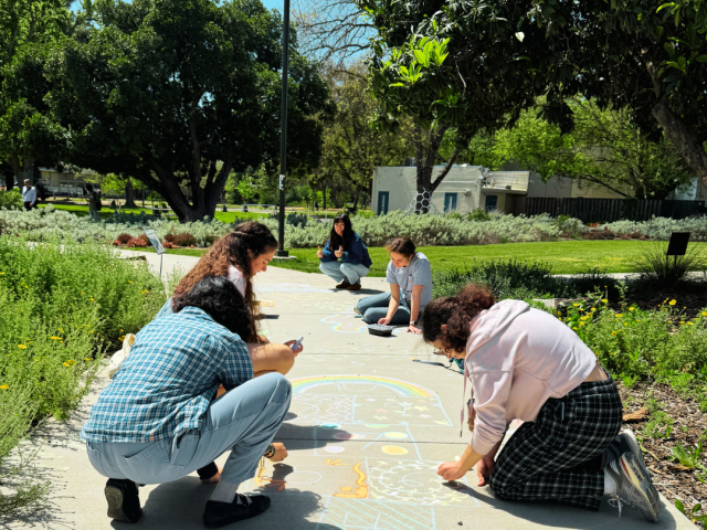 A picture of an Aggie Arts Committee event where students are using chalk to draw on the pavement outside of Cruess Hall